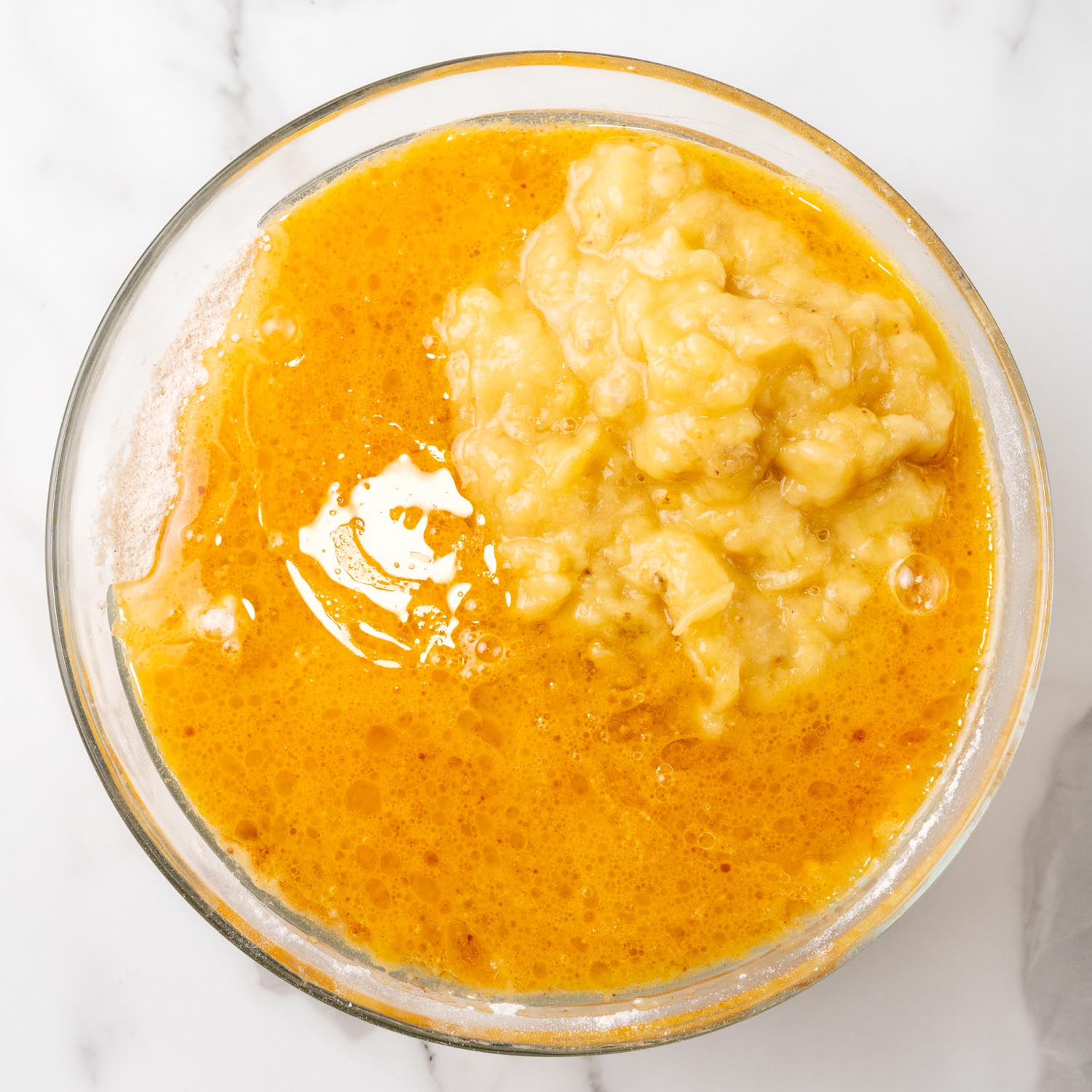 mashed banana added to oil and eggs in a glass bowl.