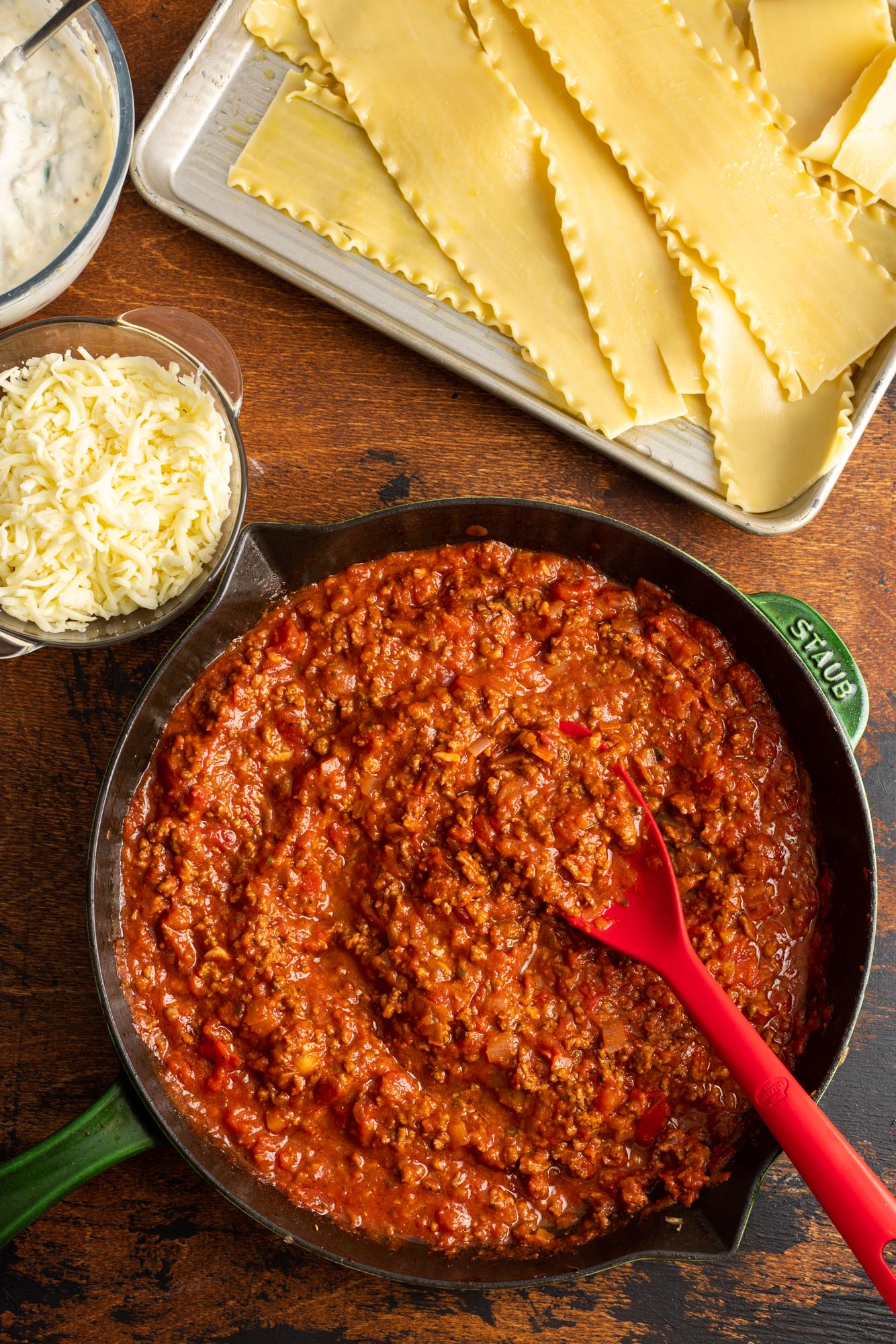 a skillet of meat sauce with a tray of cooked lasagna noodles, next to bowls of ricotta and shredded mozzarella cheese.