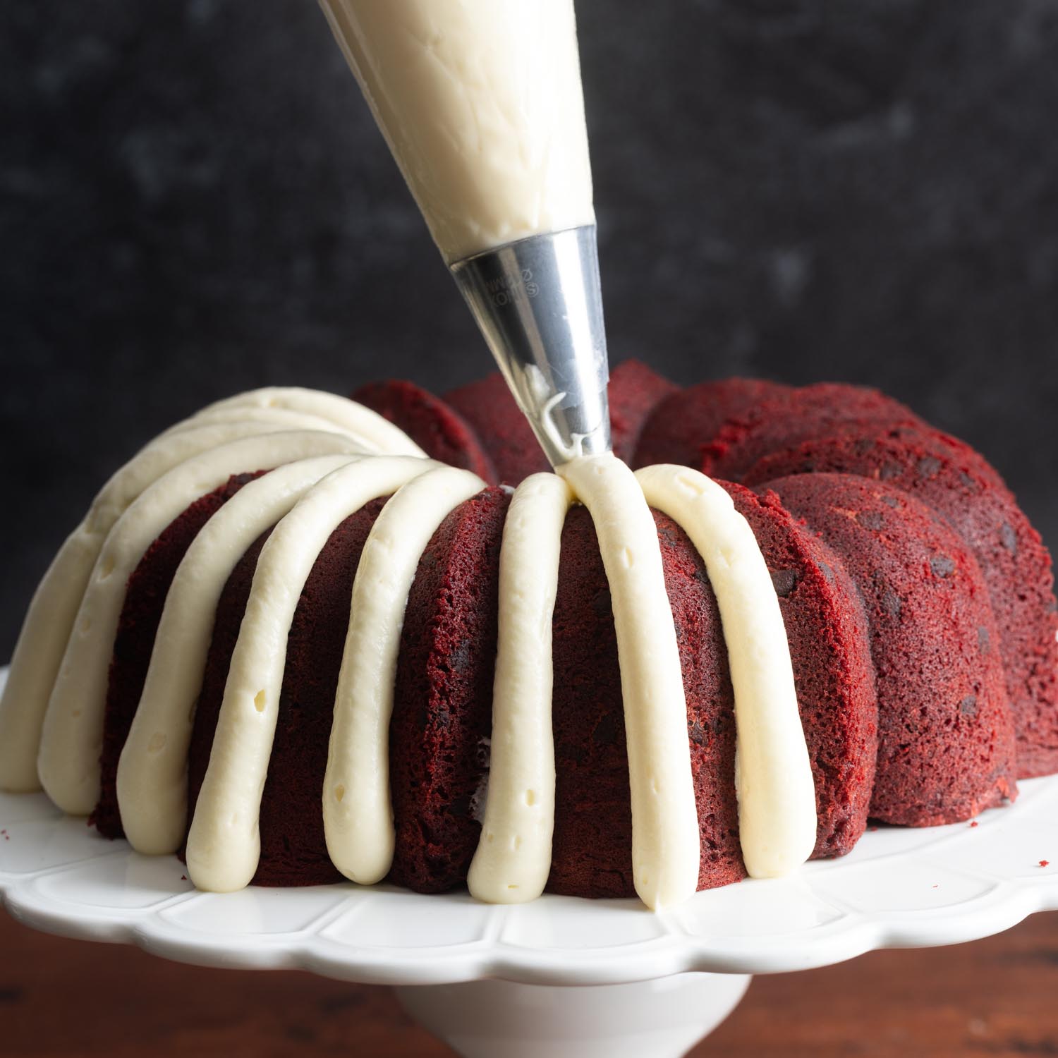 a piping bag adding thick stripes of cream cheese frosting to a red velvet bundt cake.