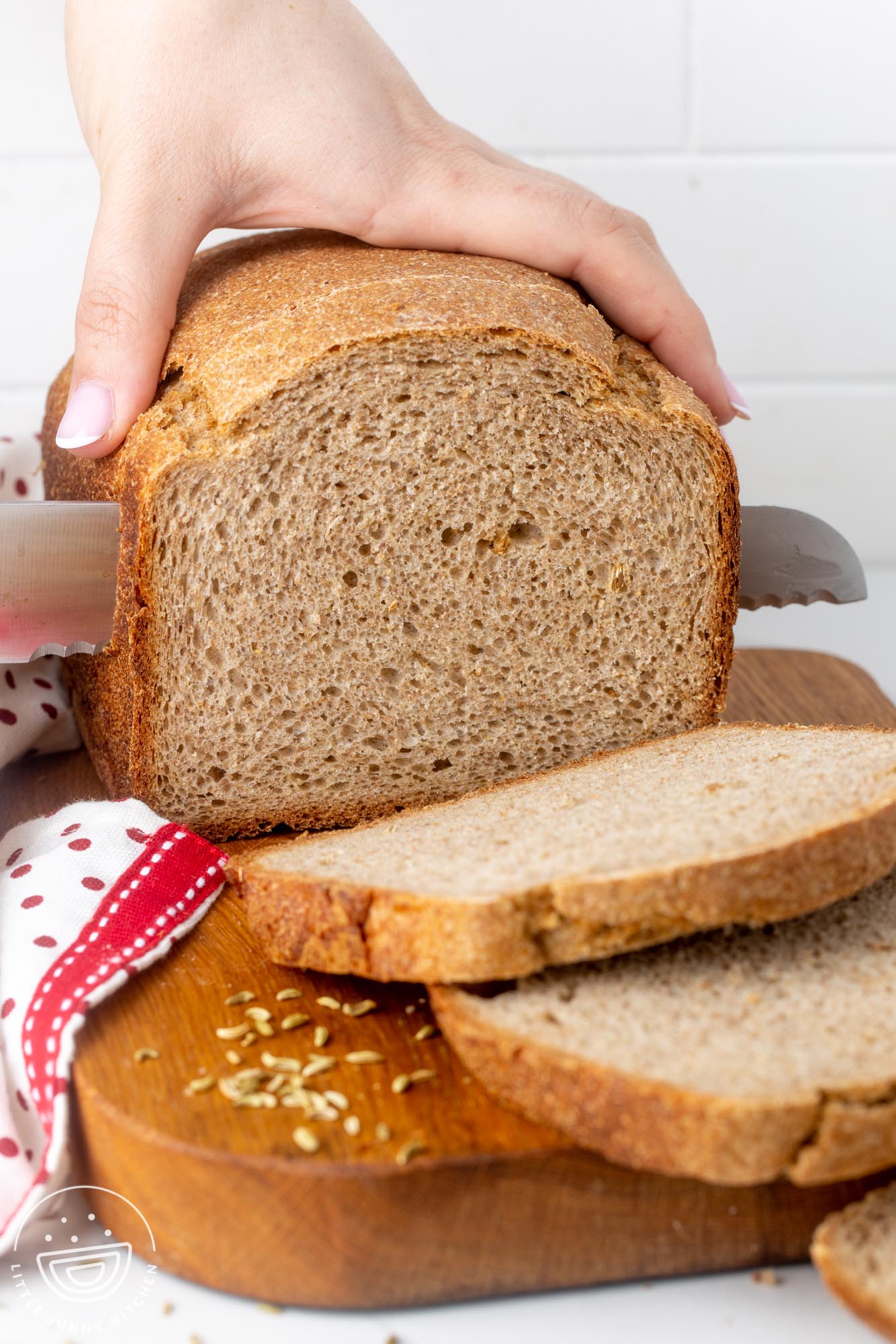 a loaf of rye bread on a wooden board. A hand is holding the loaf and cutting it into slices with a large bread knife.