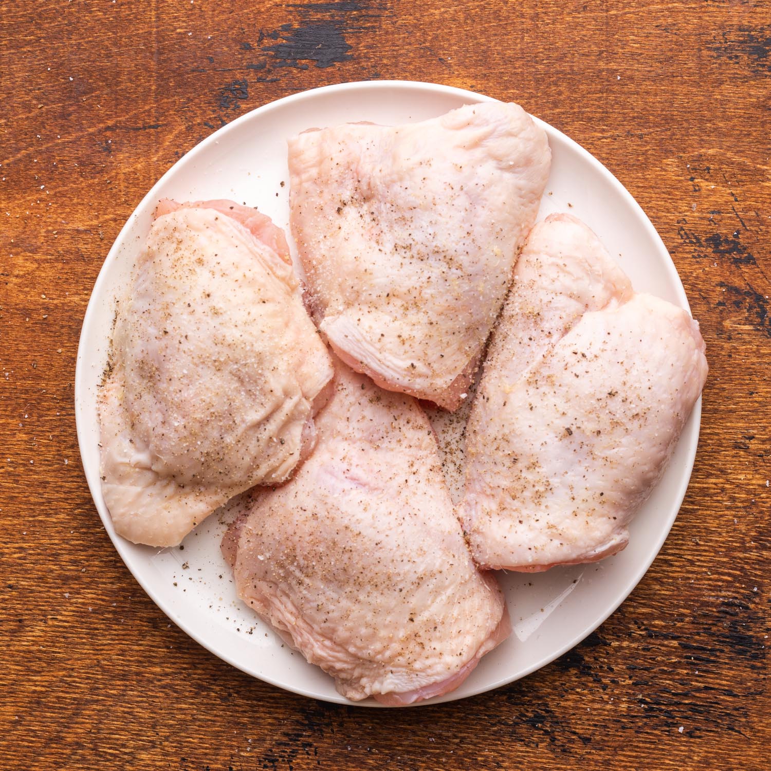 four raw bone-in chicken thighs on a white plate, seasoned with salt and pepper. The plate rests on a rustic wooden surface.