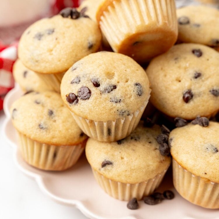 a scalloped white plate holding stacked mini muffins with chocolate chips. in the background, out of focus, is a glass of milk and a red and white checked towel.