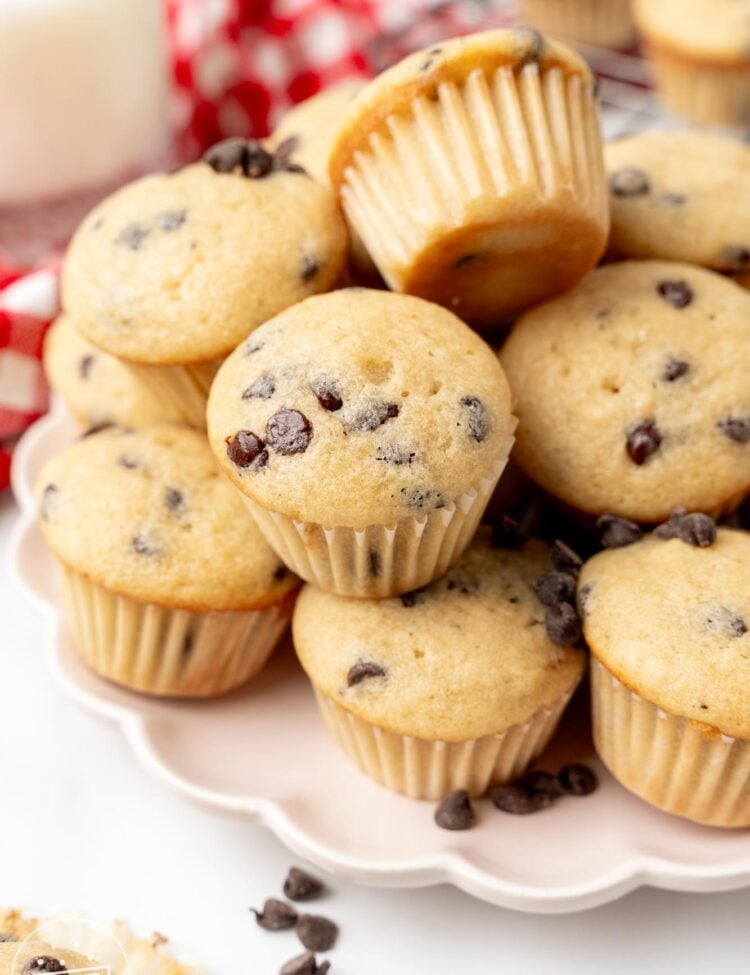 a scalloped white plate holding stacked mini muffins with chocolate chips. in the background, out of focus, is a glass of milk and a red and white checked towel.