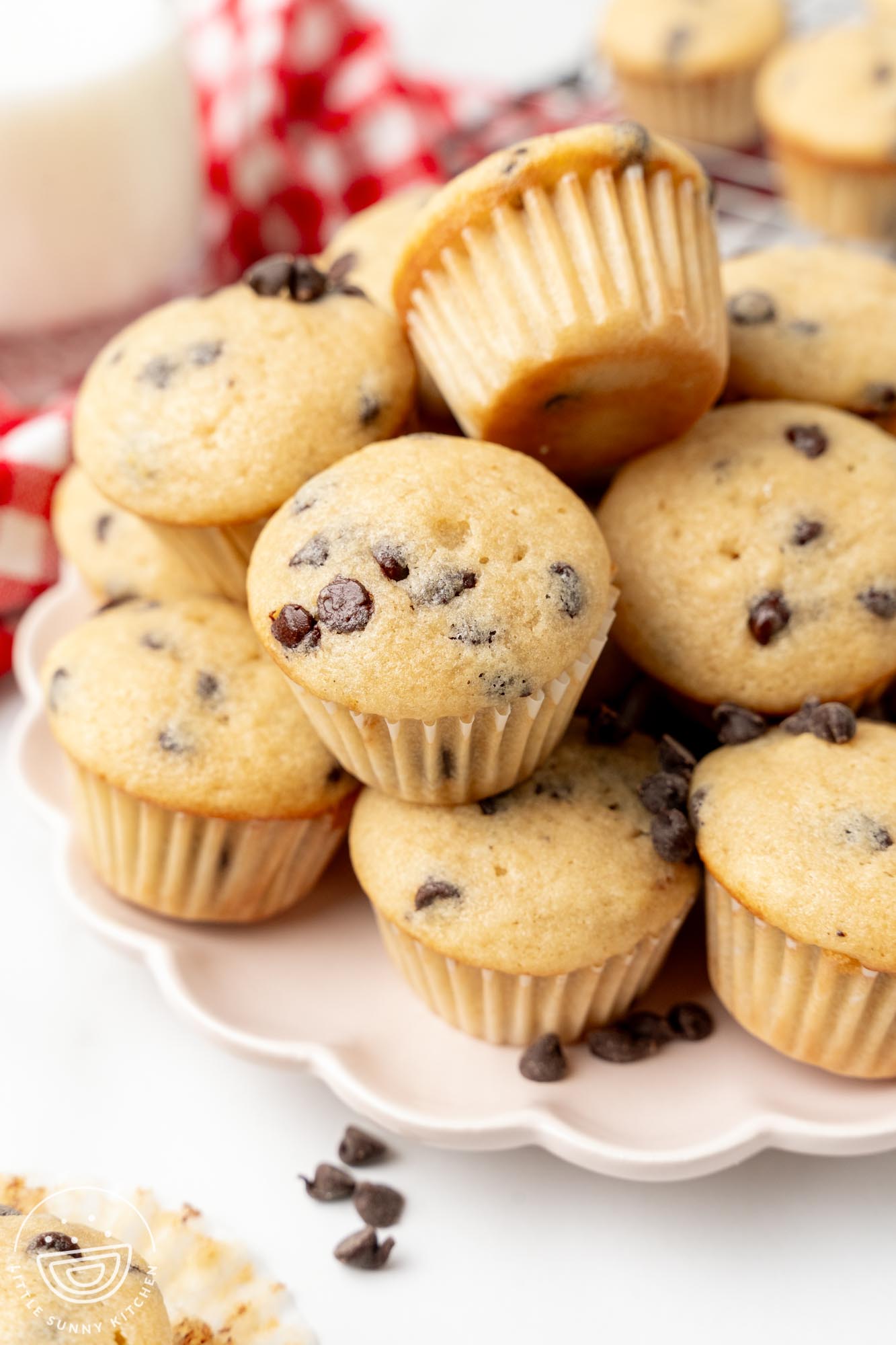 a scalloped white plate holding stacked mini muffins with chocolate chips. in the background, out of focus, is a glass of milk and a red and white checked towel.