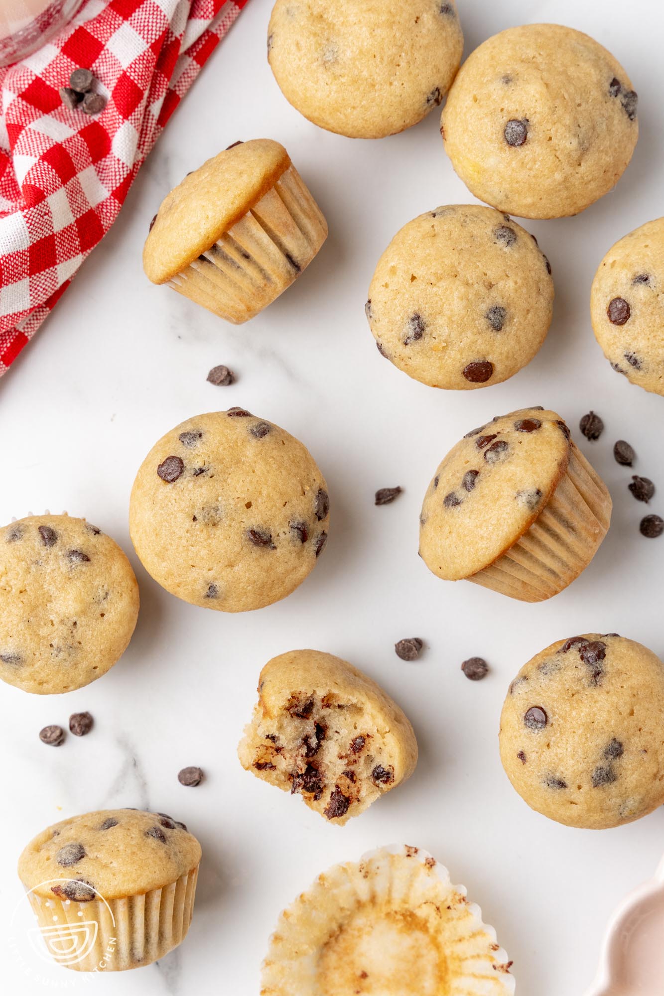 mini chocolate chip muffins spread out on a marble counter next to a red and white checked towel.