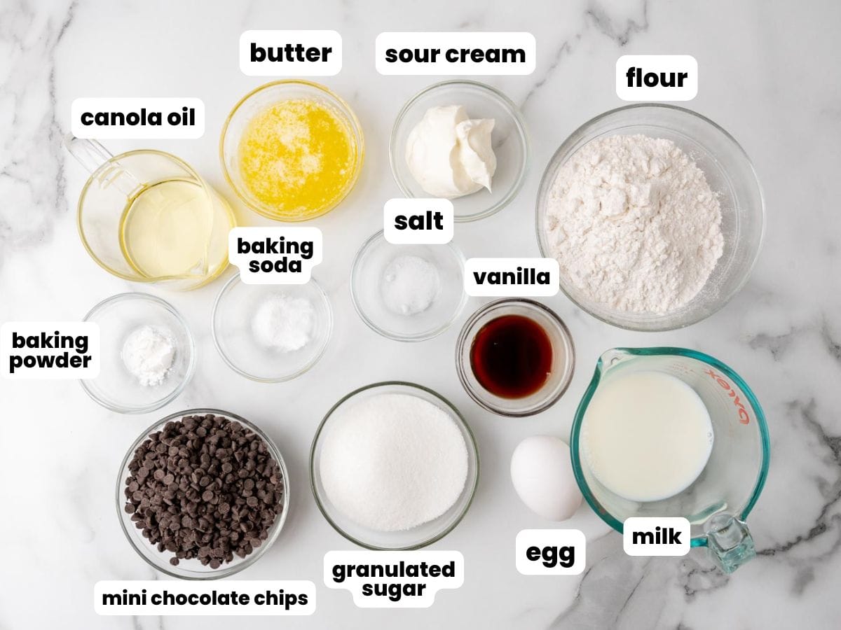 Baking ingredients for mini chocolate chip muffins in small glass bowls, arranged on a marble surface.