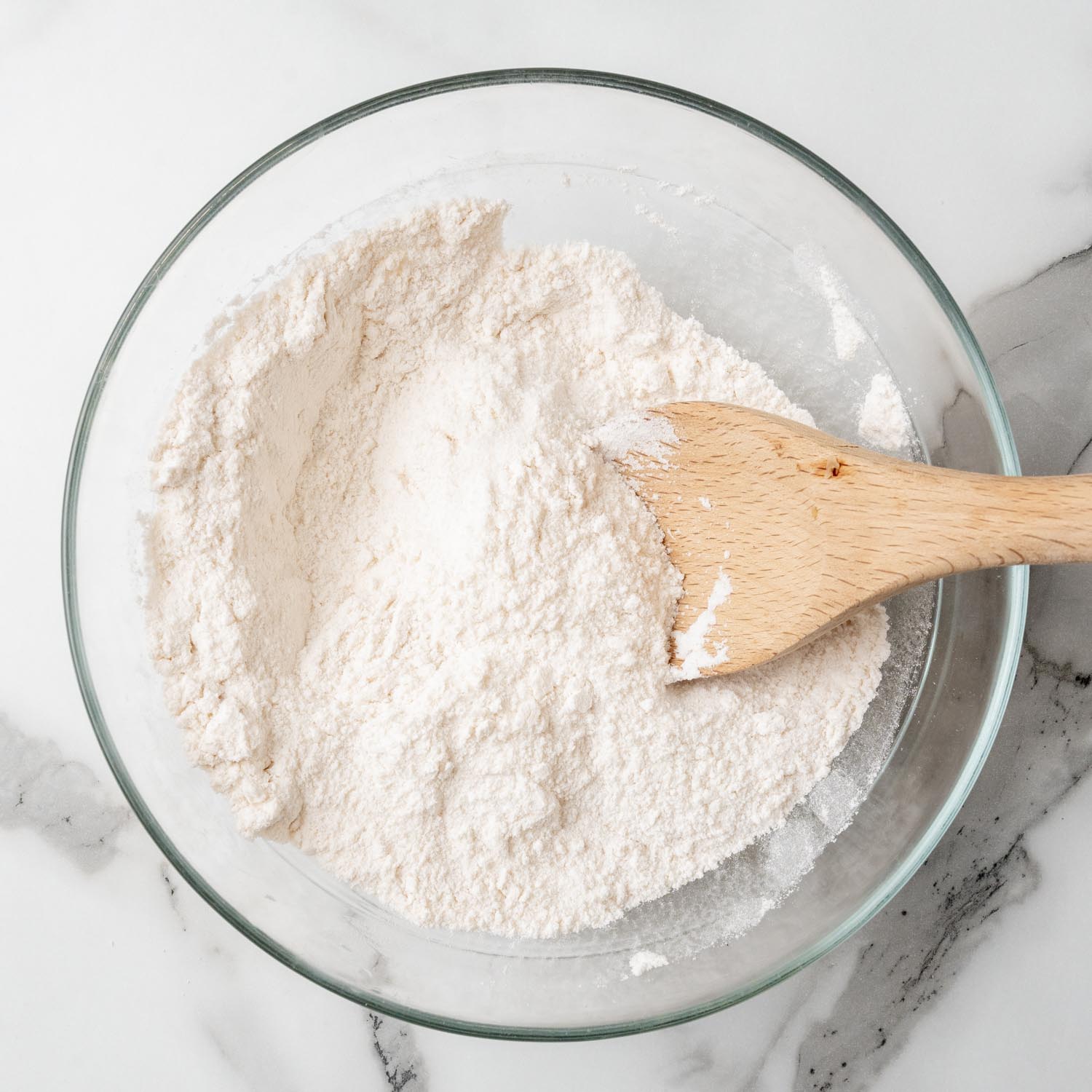 A glass mixing bowl of flour. There is a wooden spoon in the bowl and it is sitting on a marble surface.