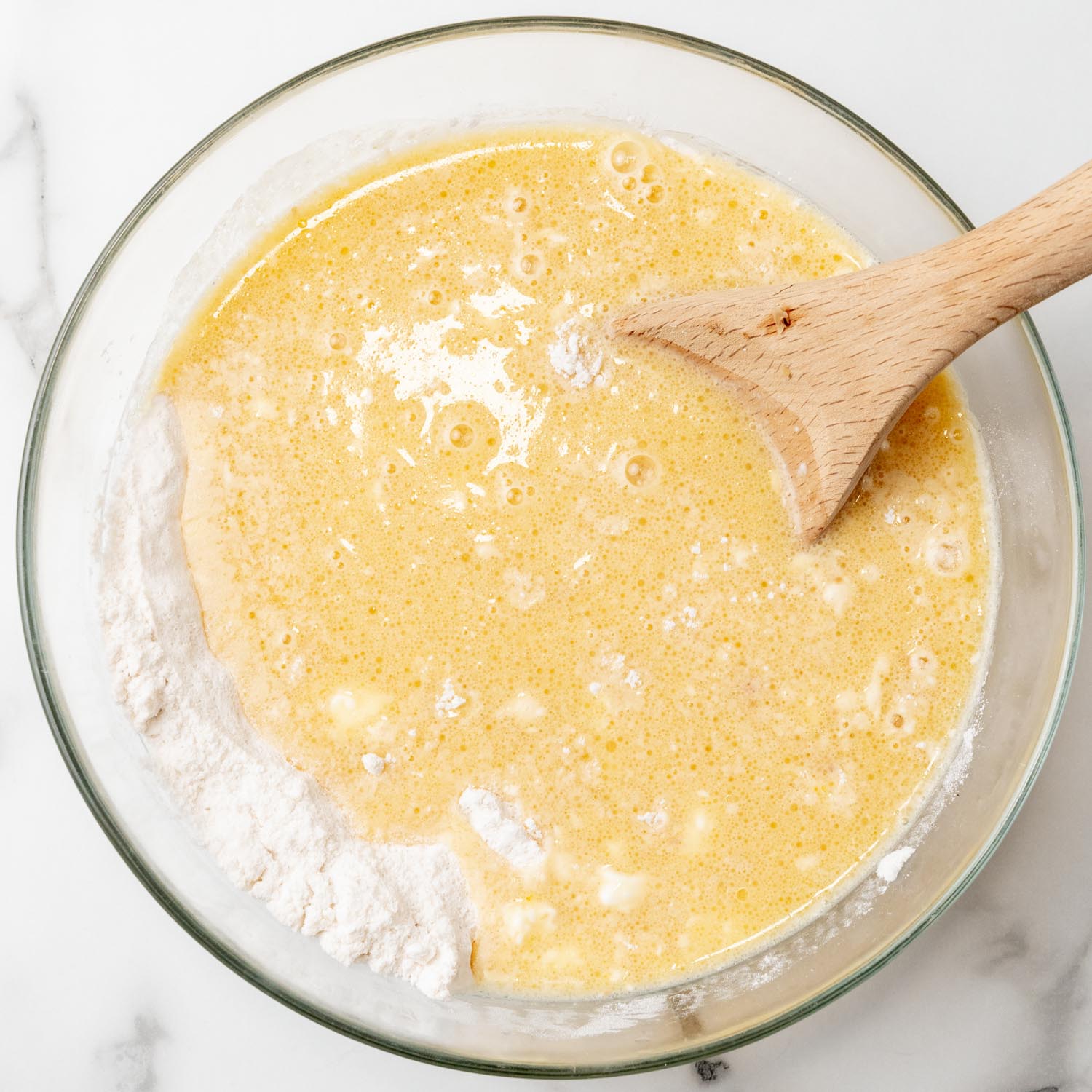 Wet ingredients added to dry ingredients for muffin batter in a glass mixing bowl, stirred with a large wooden spoon.