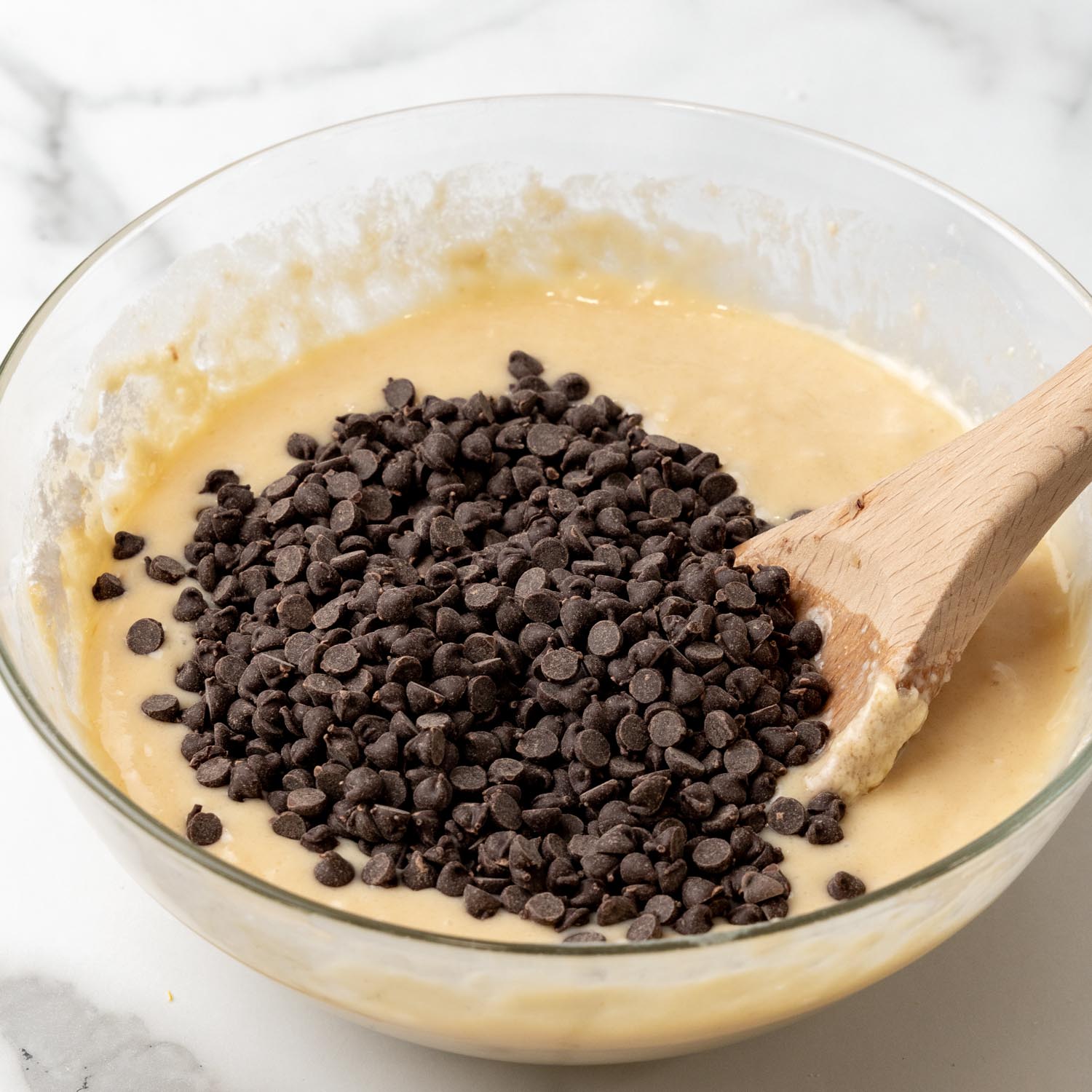 Mini chocolate chips added to a glass bowl of muffin batter that is sitting on a marble surface.