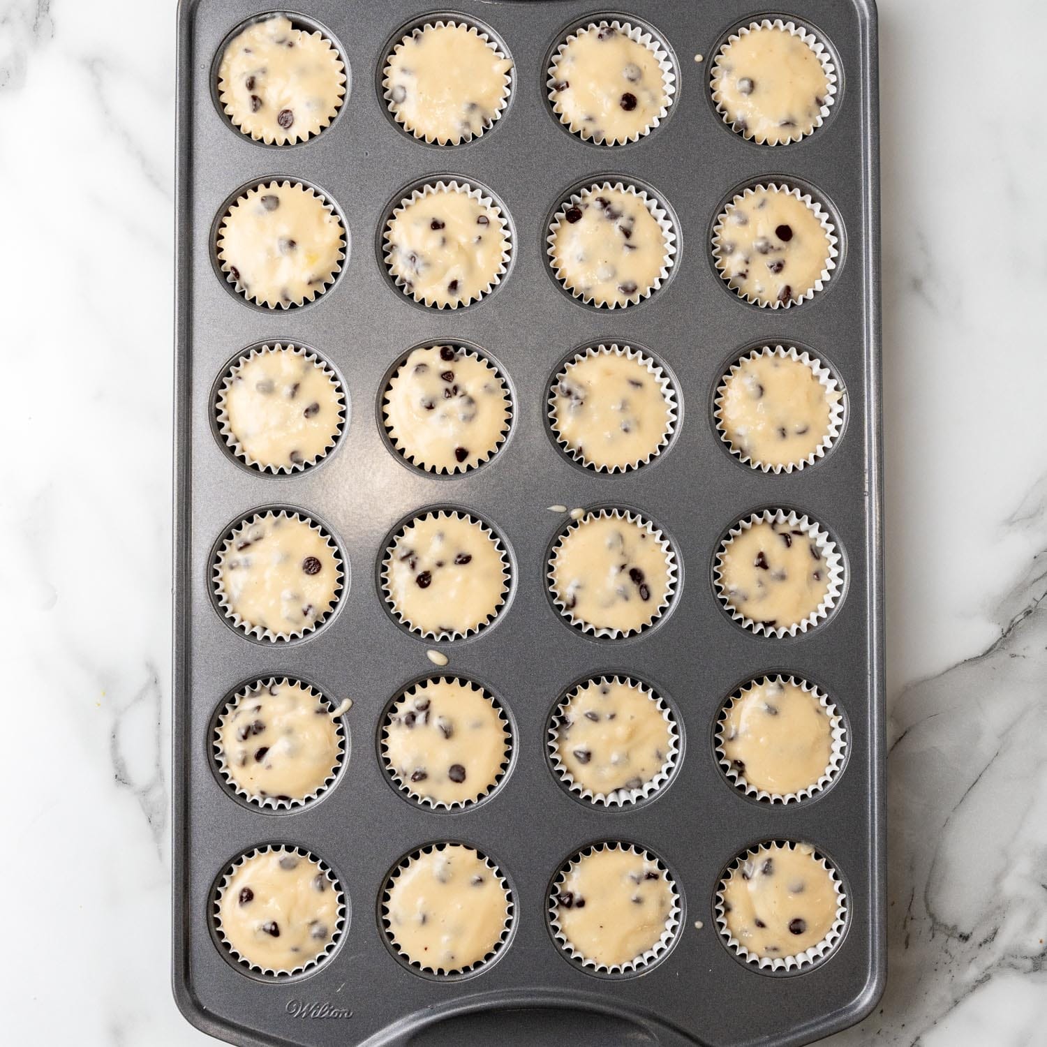 overhead shot of a 24 space mini muffin tin lined with papers that are filled with chocolate chip muffin batter.