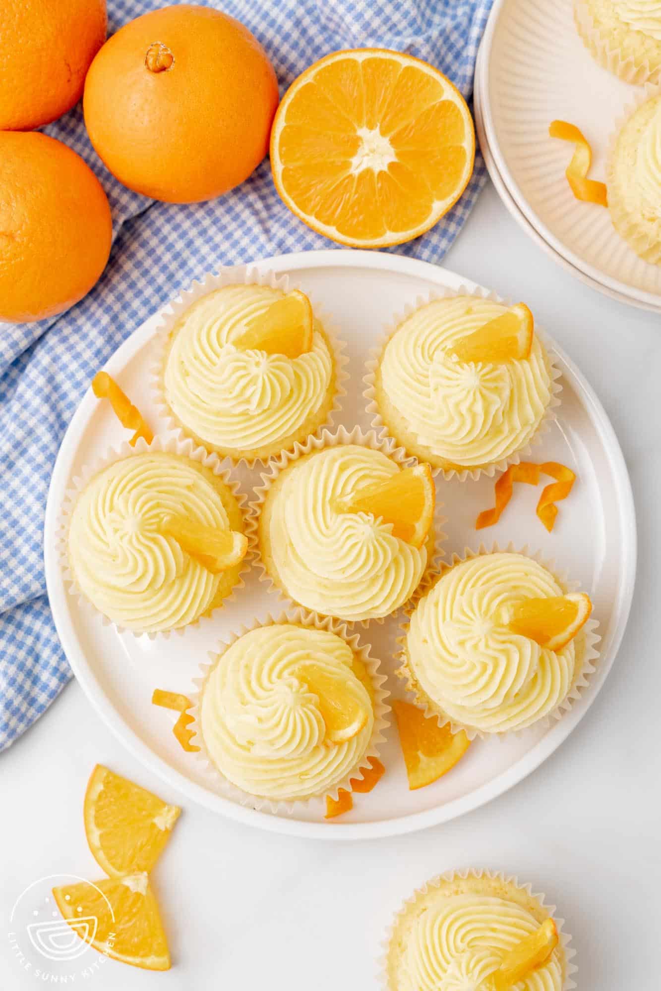 overhead view of a plate of 6 orange cupcakes with frosting and orange wedge decorations. Next to the plate are fresh oranges and curls of orange peel.