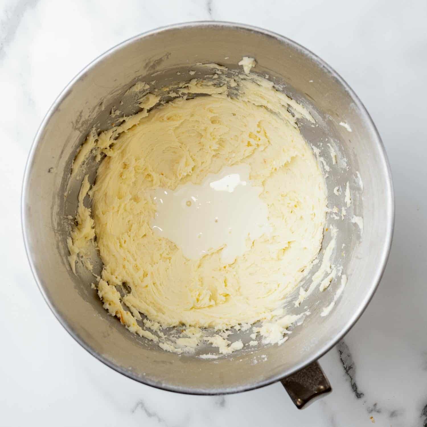 heavy cream added to orange buttercream in a metal mixing bowl on a marble counter.