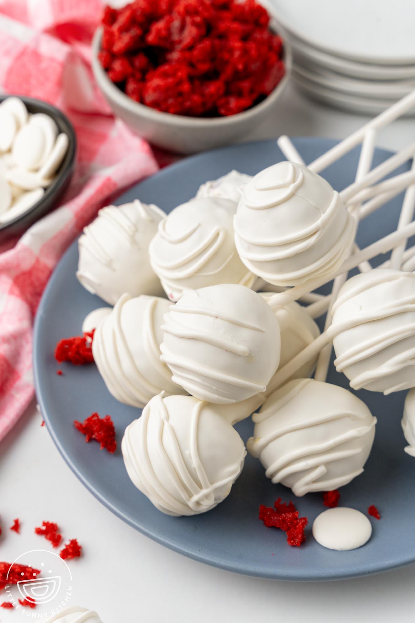 red velvet cake pops coated in white chocolate with drizzle on a blue plate. Crumbs of red velvet cake are around the pops.