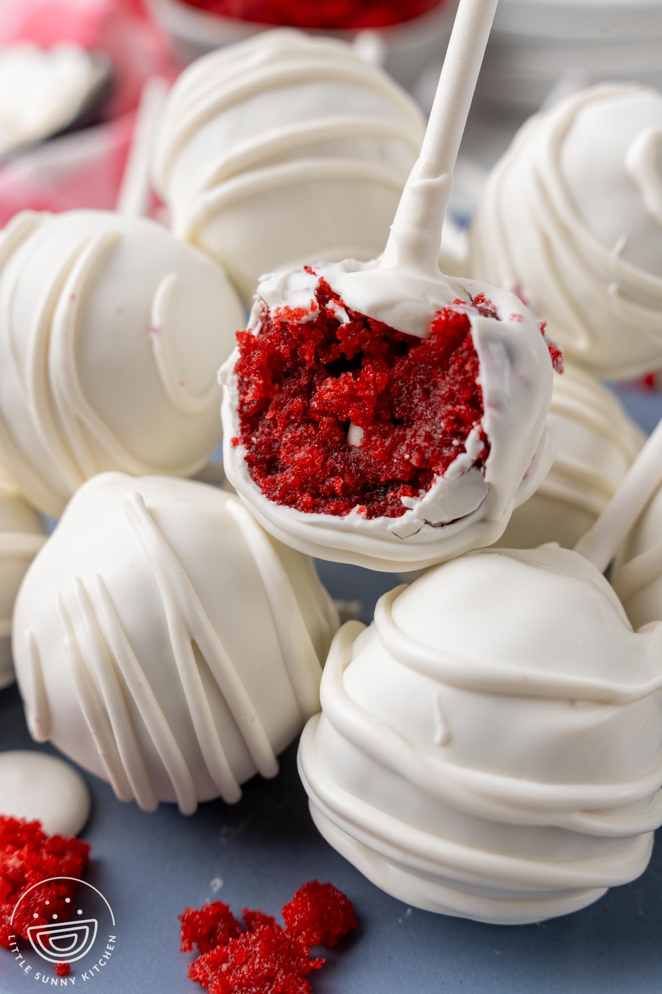 White coated cake pops on a plate. One on top has a bite taken to show the red velvet cake inside.