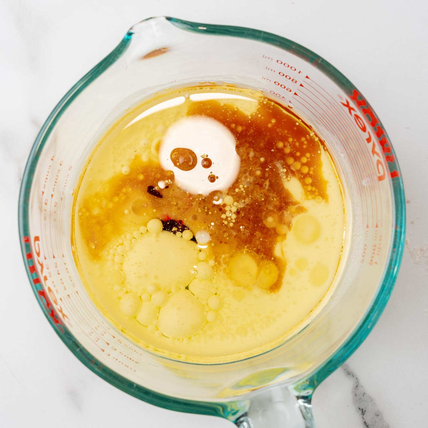 buttermilk, oil, and vinegar added to wet ingredients for red velvet cake in a pyrex measuring cup. The mixture has bubbles and cells from the oil.