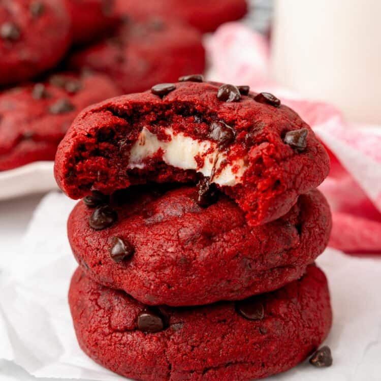 a stack of three red velvet cream cheese cookies with chocolate chips. The top cookie has a bite taken to show the gooey cream cheese filling inside.