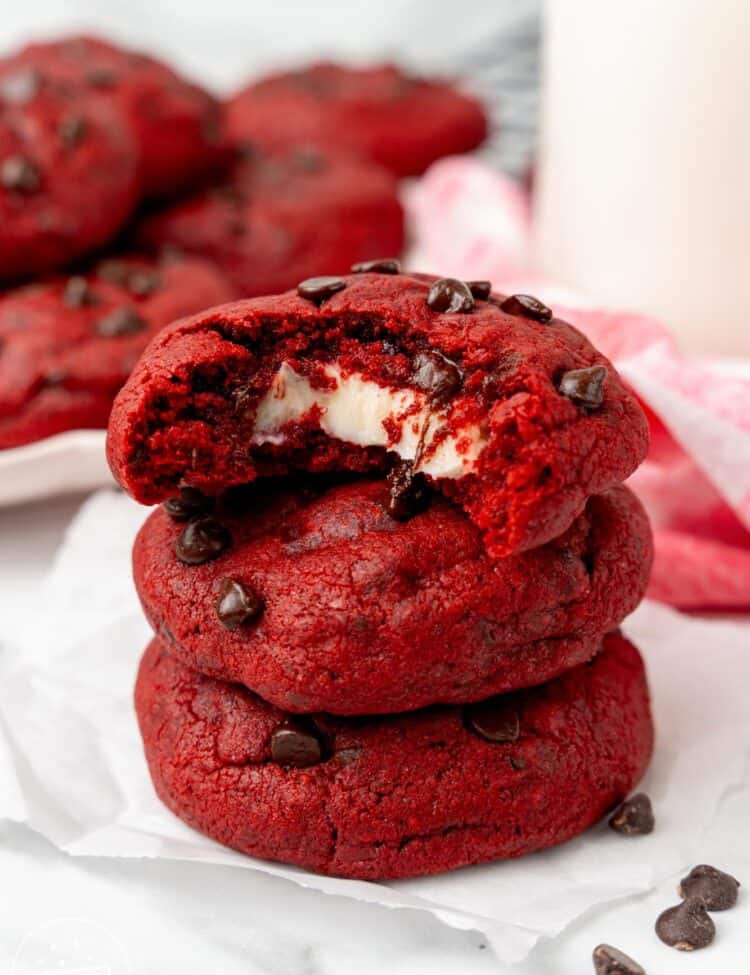 a stack of three red velvet cream cheese cookies with chocolate chips. The top cookie has a bite taken to show the gooey cream cheese filling inside.