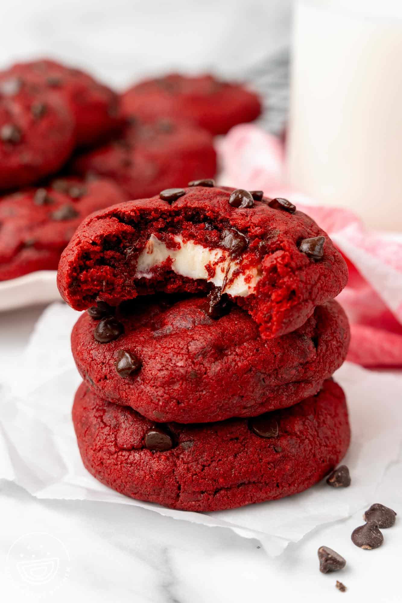 a stack of three red velvet cream cheese cookies with chocolate chips. The top cookie has a bite taken to show the gooey cream cheese filling inside.