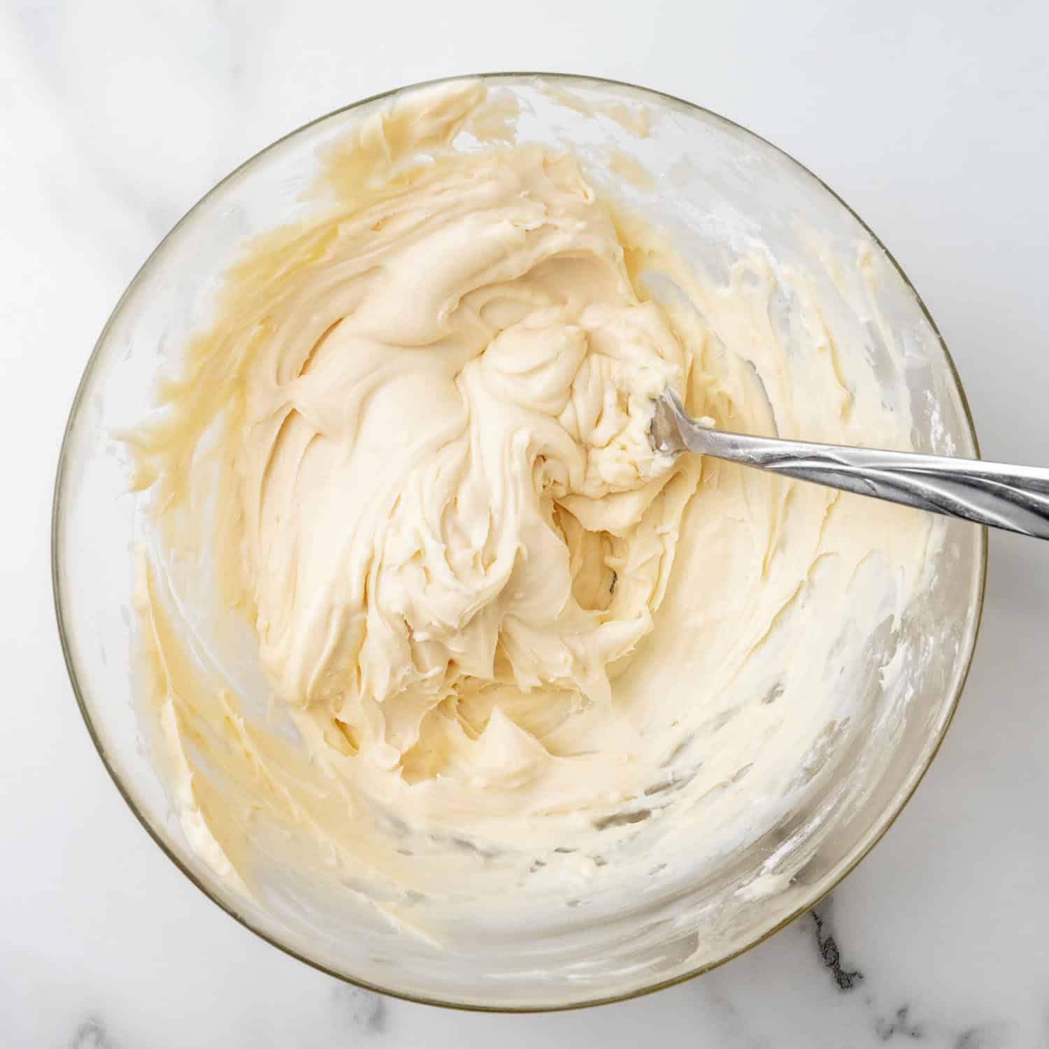 Cream cheese filling for red velvet cookies in a glass mixing bowl stirred with a metal spoon.