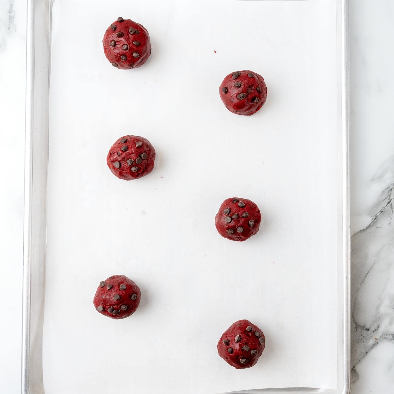 large stuffed balls of red velvet cookie dough with mini chocolate chips on the outside. The dough balls are arranged far apart on a parchment lined baking sheet.