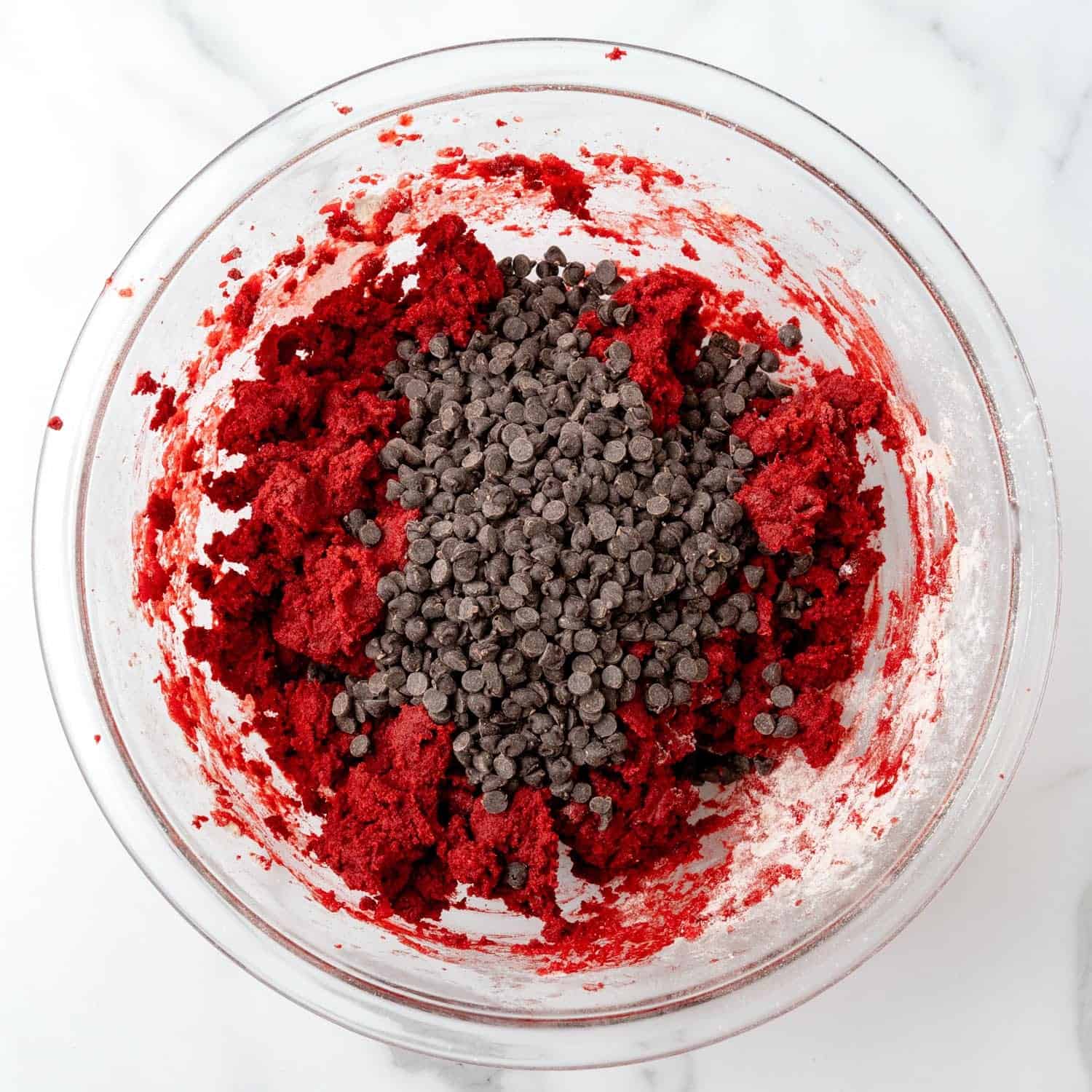 mini chocolate chips added to deep red velvet cookie dough in a glass mixing bowl set on a marble counter.