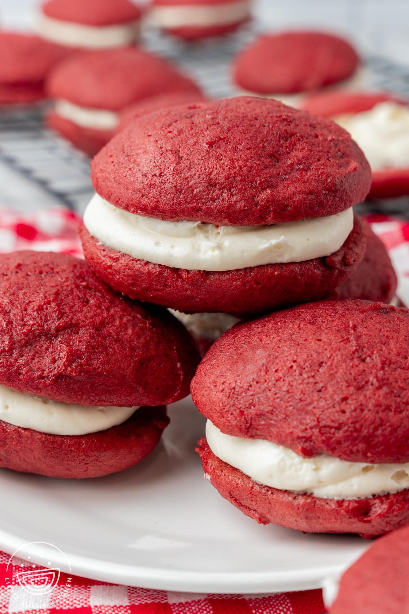 a white plate on a red and white checked towel. On the plate is a stack of four red velvet whoopie pies with cream cheese filling.