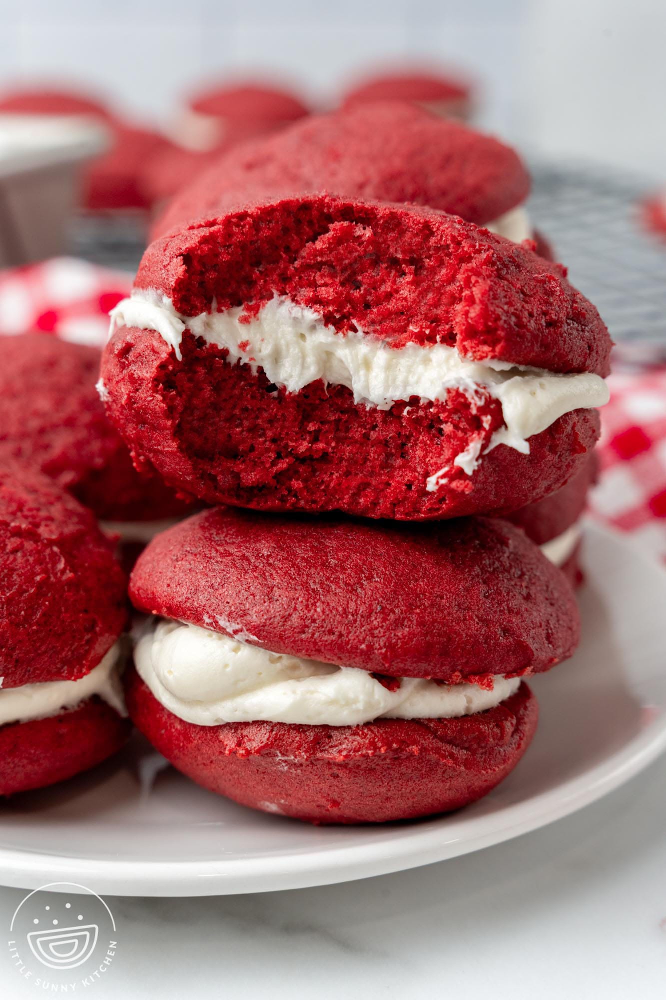 red velvet whoopie pie sandwiches on a white plate. One cookie has a bite taken to show the fluffy interior and creamy white frosting.