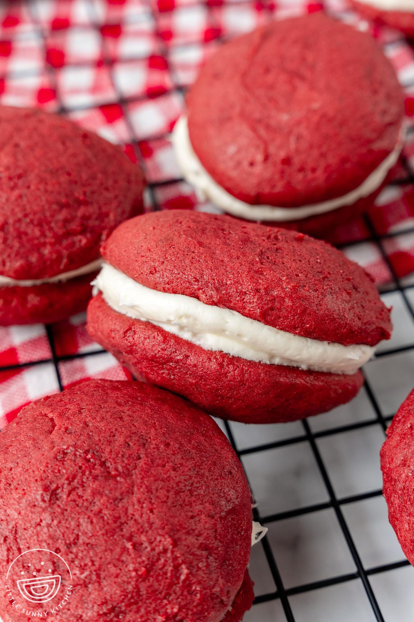 red velvet whoopie pies on a metal cooling rack on top of a red and white checked towel.