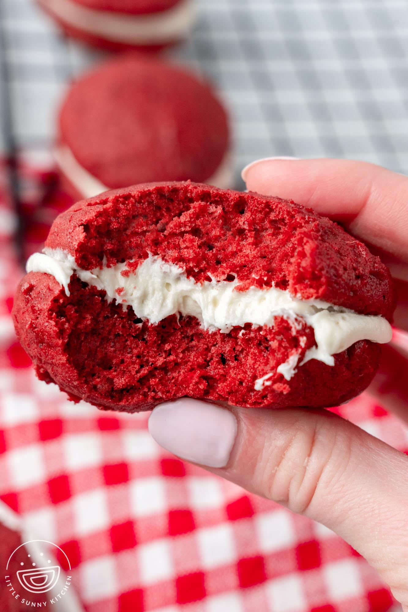 a hand holding a red velvet whoopie pie with a bite taken to show the fluffy texture and creamy filling.