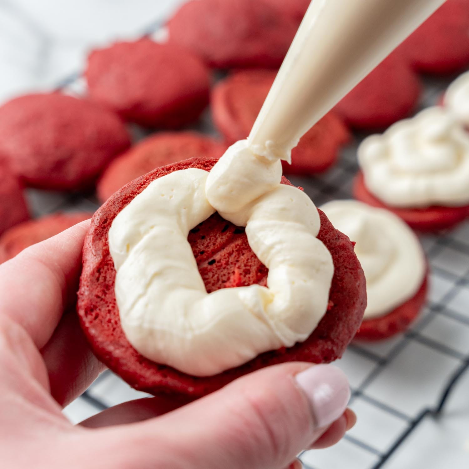 a hand holding one half of a red velvet whoopie pie while piping cream cheese icing onto it.