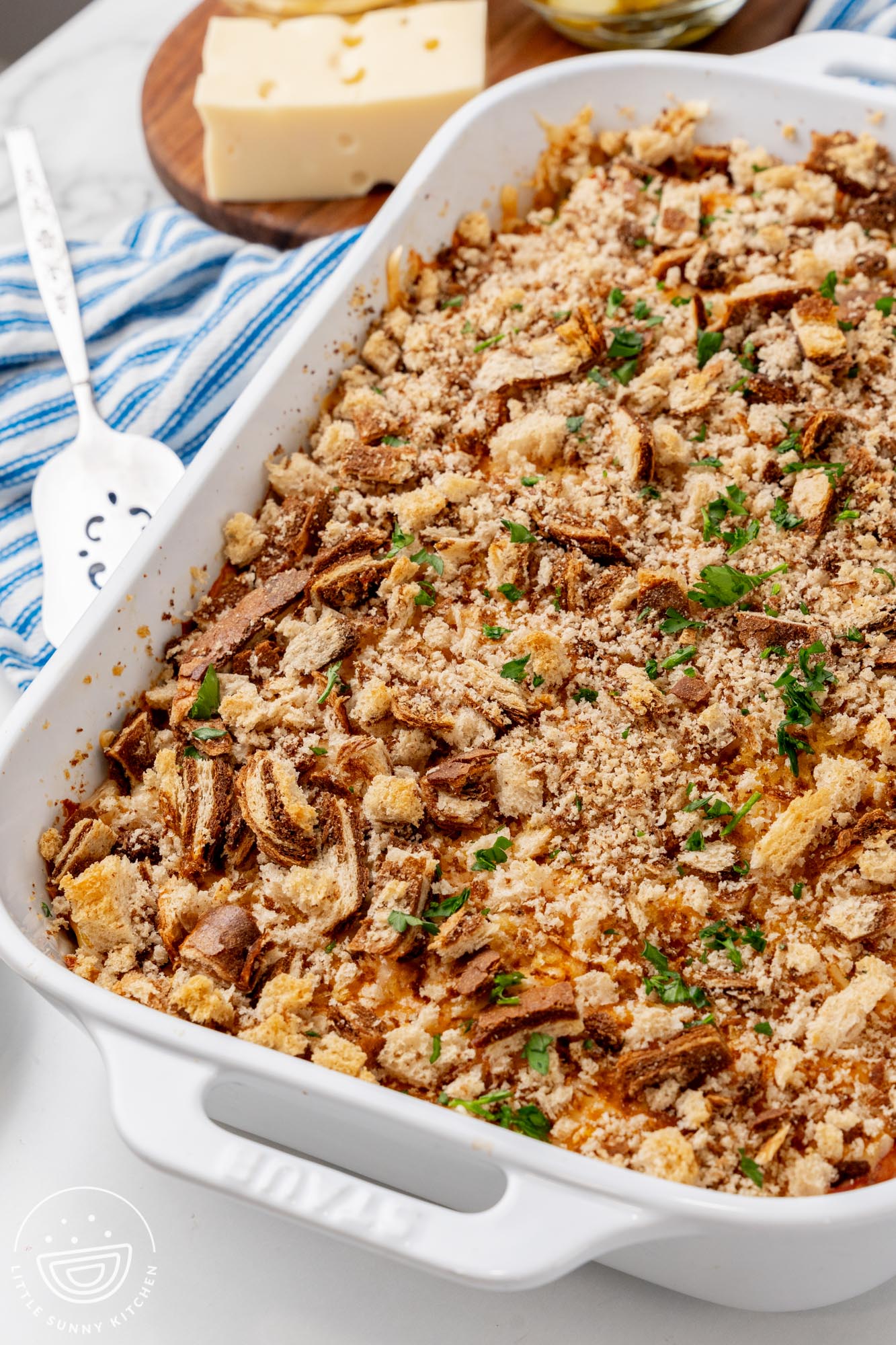 A white rectangular casserole dish of Reuben Casserole topped with marble rye bread crumbs and chopped parsley. Next to the pan is a serving spoon and a piece of swiss cheese.