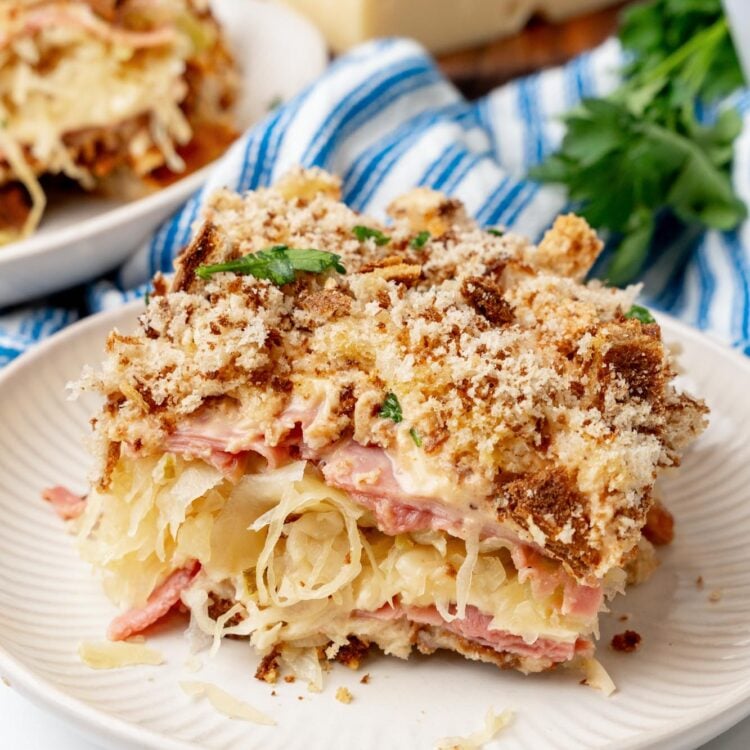 A square slice of reuben casserole on a white plate. There are layers of sliced corned beef, sauerkraut, swiss cheese, and rye breadcrumbs.