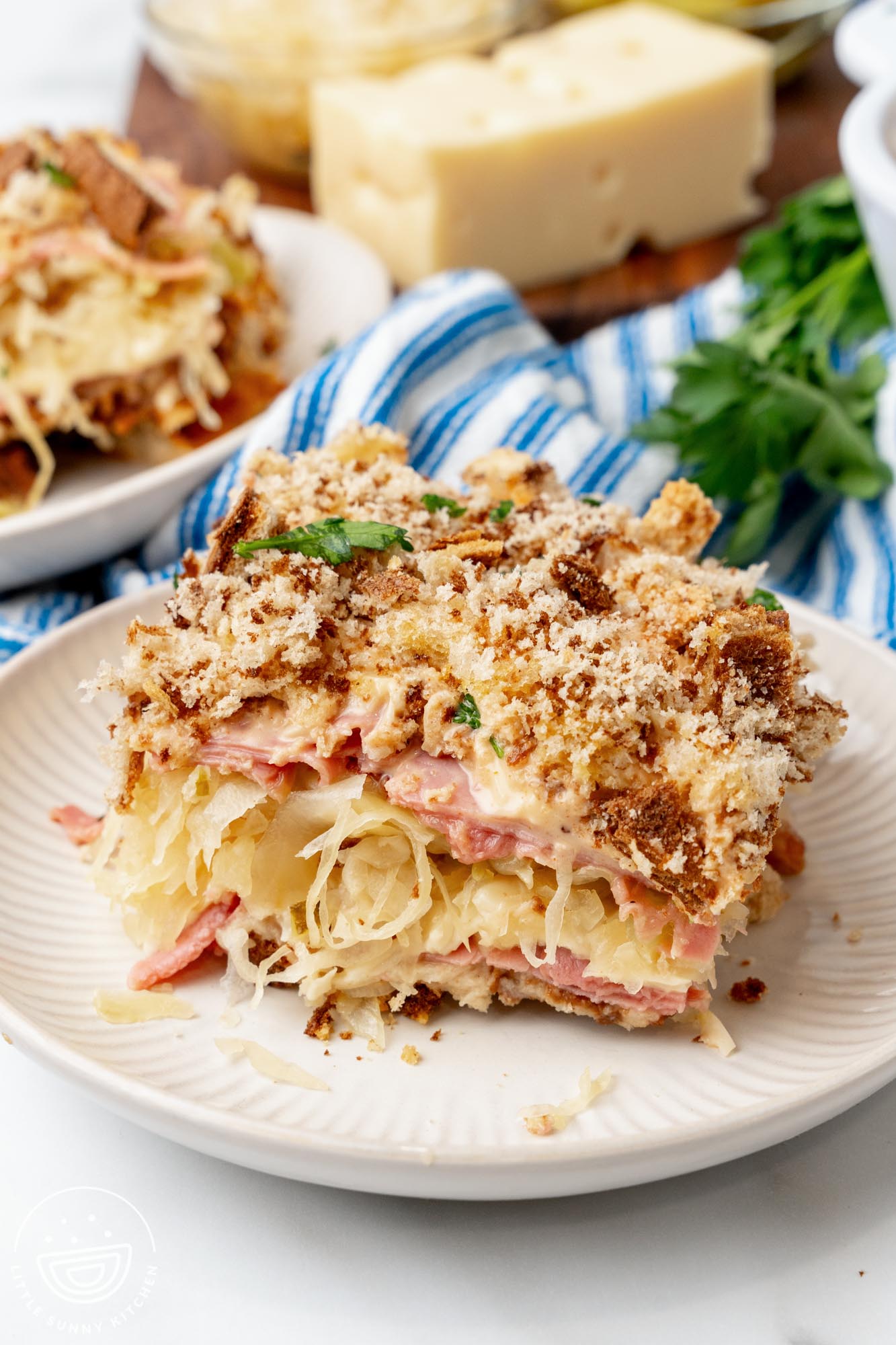 A square slice of reuben casserole on a white plate. There are layers of sliced corned beef, sauerkraut, swiss cheese, and rye breadcrumbs.