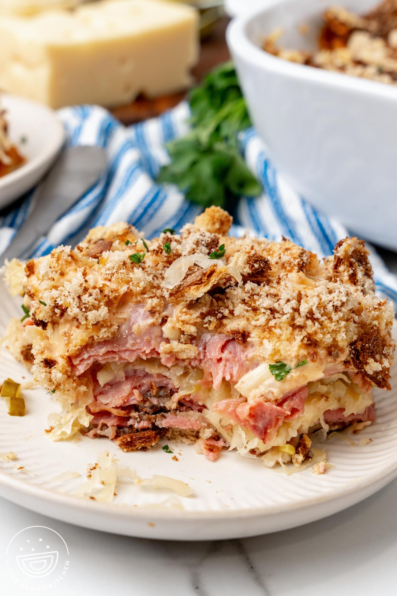 A serving of layered Reuben sandwich casserole with corned beef, swiss cheese, and sauerkraut, topped with rye breadcrumbs, on a small white plate.