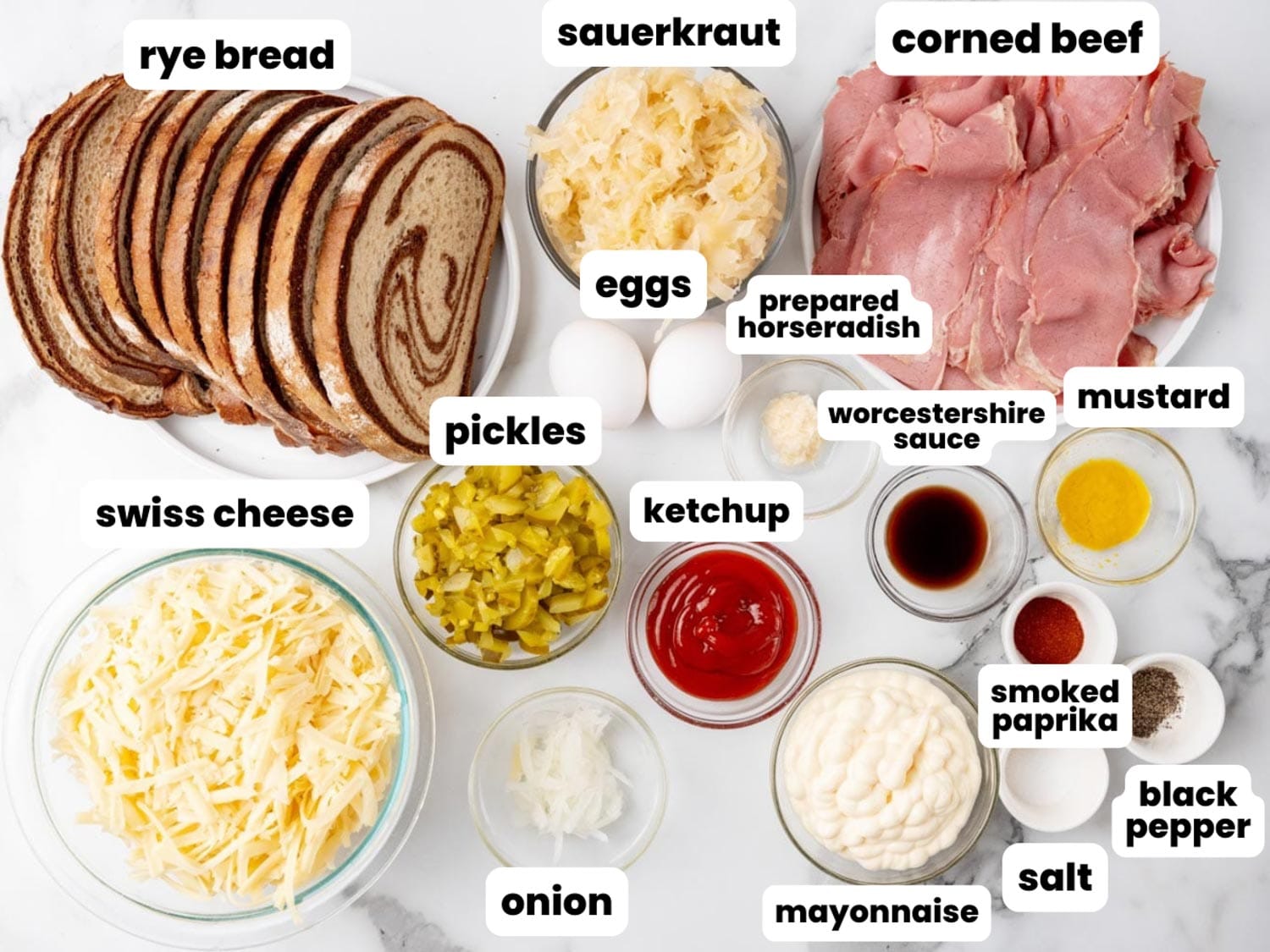 Ingredients for Reuben Casserole in small bowls on a marble surface, including deli corned beef, rye bread, swiss cheese, and sauerkraut.