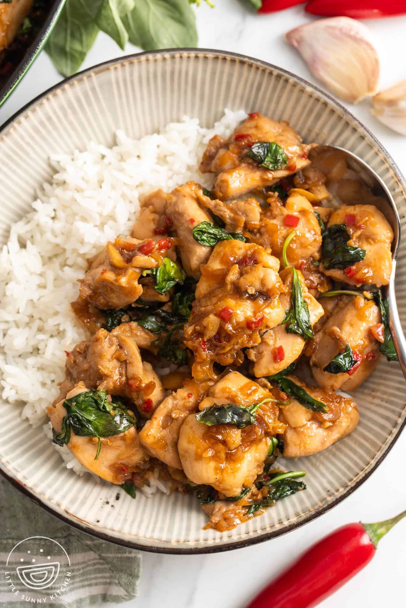 a ceramic bowl of rice topped with thai basil chicken. Around the bowl are chili peppers, garlic cloves and fresh thai basil.