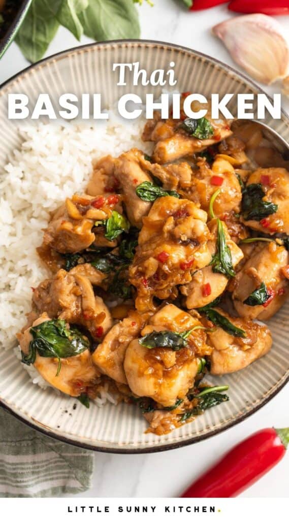 a ceramic bowl of rice topped with thai basil chicken. Around the bowl are chili peppers, garlic cloves and fresh thai basil. Text overlay says "Thai Basil Chicken"