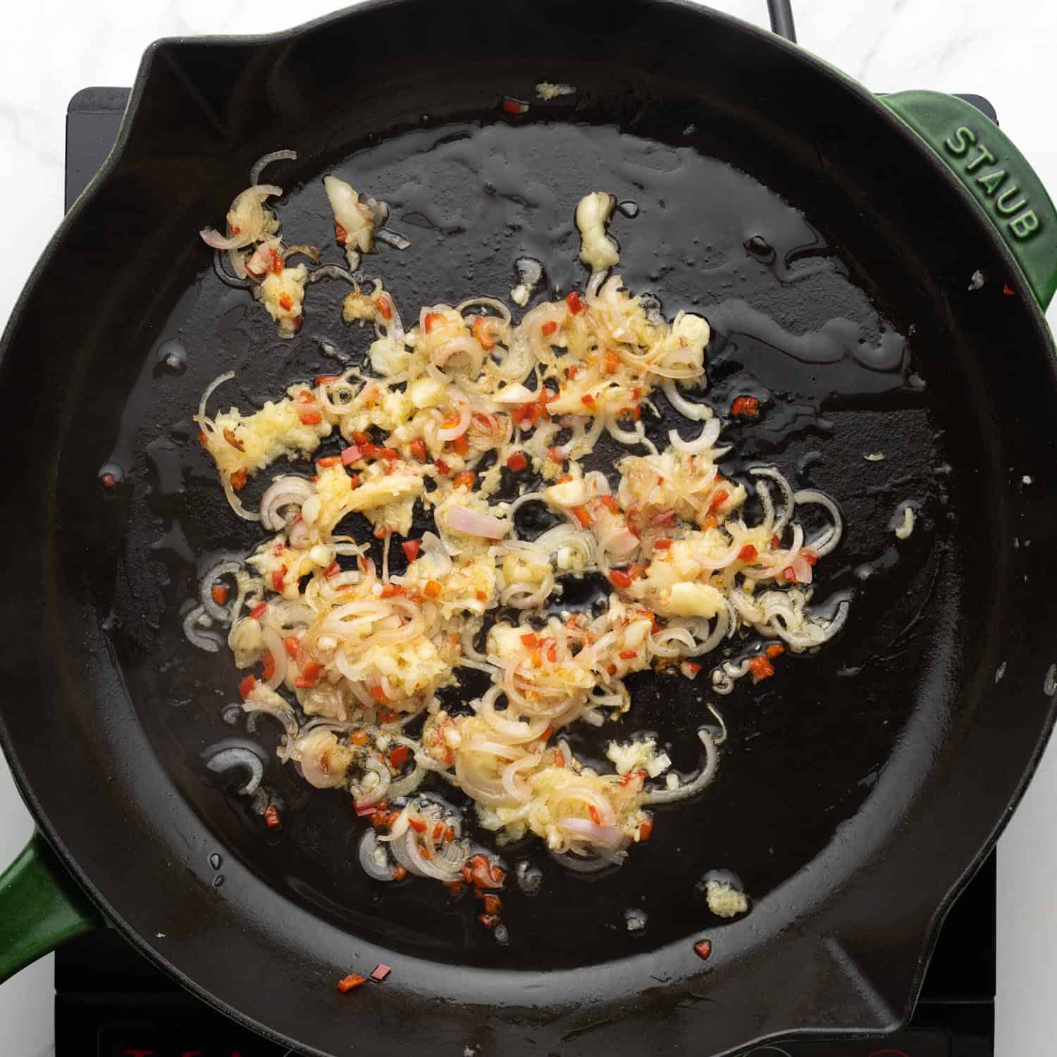 shallots, garlic, and finely diced hot peppers cooking in oil in a green Staub cast iron skillet.