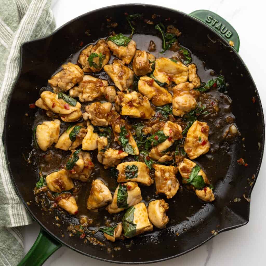 Overhead view of a skillet that has been removed from the stove, filled with cooked thai basil chicken with wilted basil leaves.