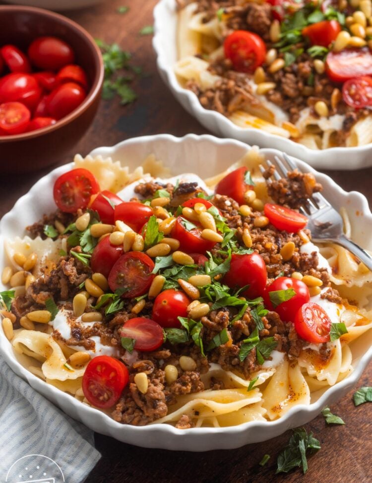 Two bowls of Turkish pasta topped with yogurt, ground beef, tomatoes, pinenuts, and herbs.