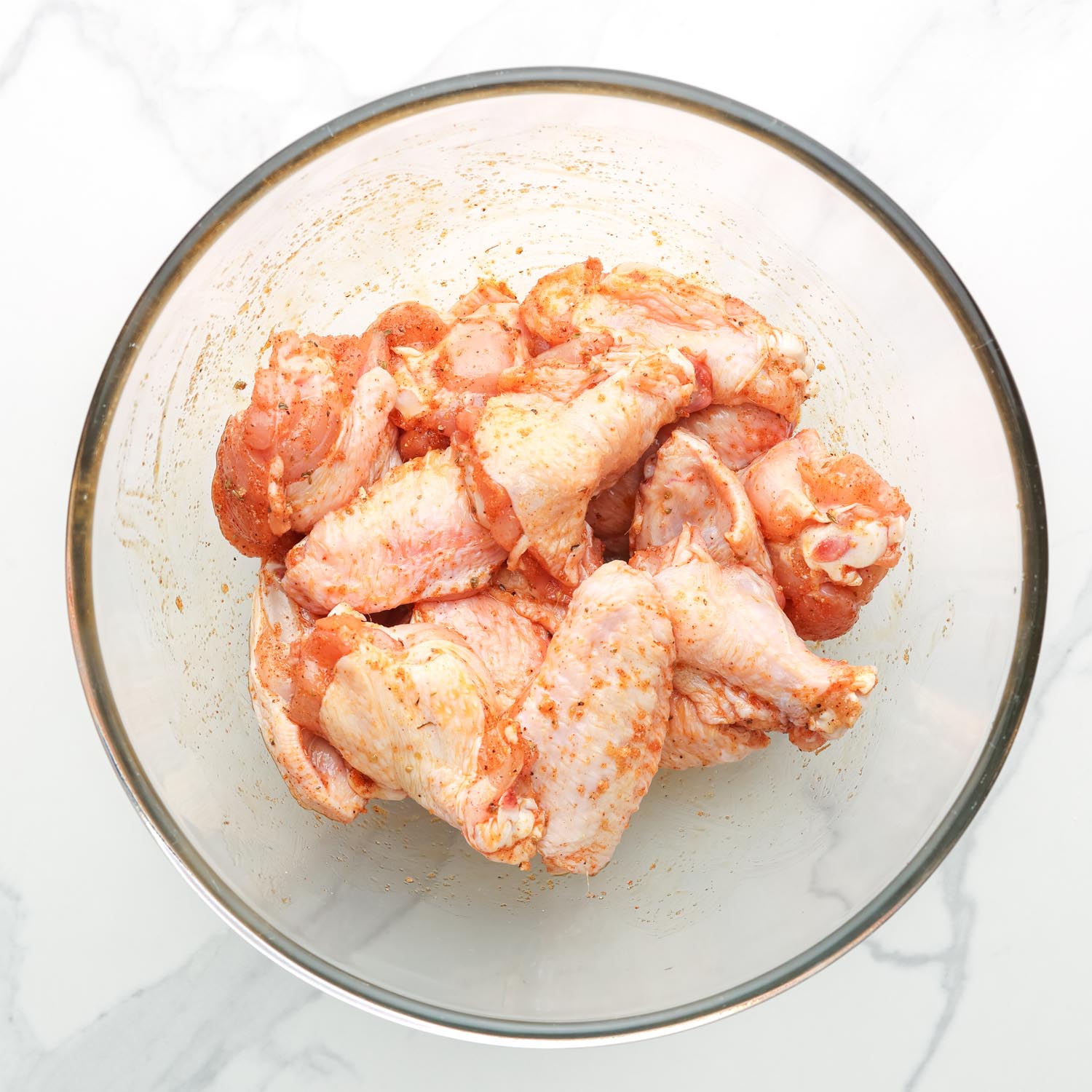 Raw chicken wings in a glass bowl tossed with oil and seasonings.