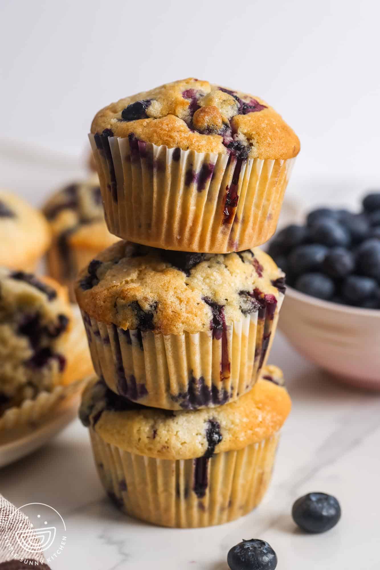 Three homemade blueberry muffins stacked on top of each other. In the background is a bowl of fresh berries and more blueberry muffins.