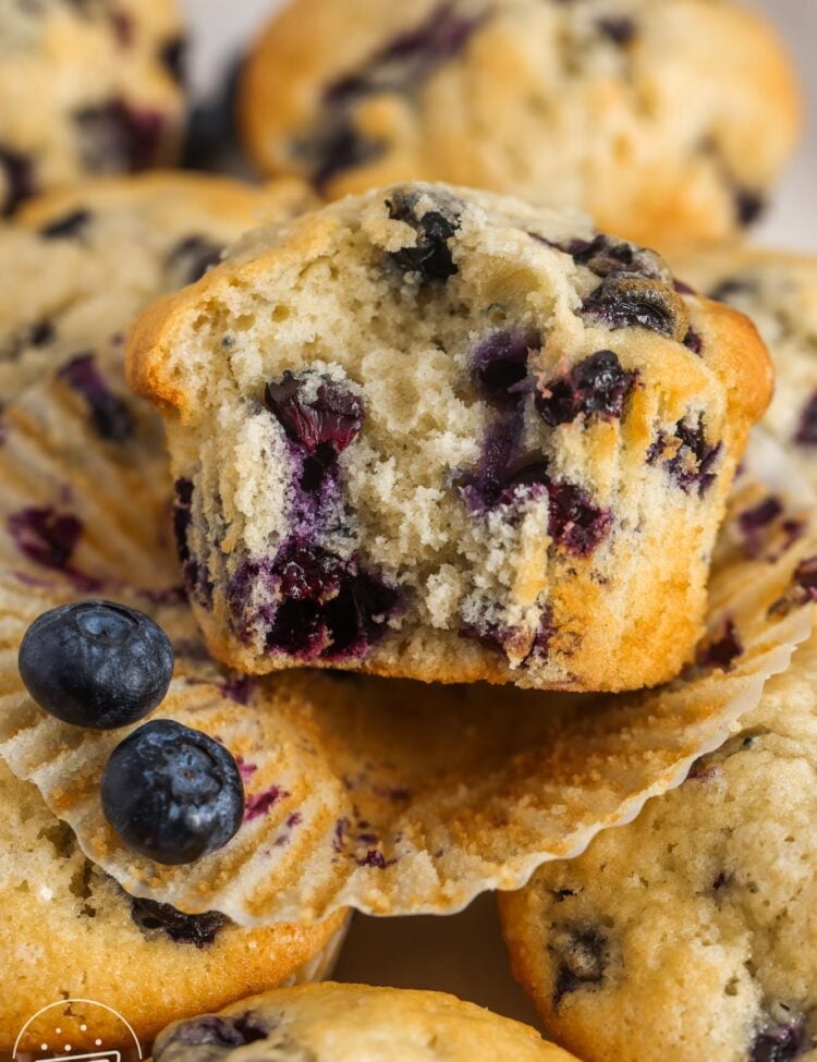 a blueberry muffin that has had the paper peeled away and a bite taken, on top of other blueberry muffins.