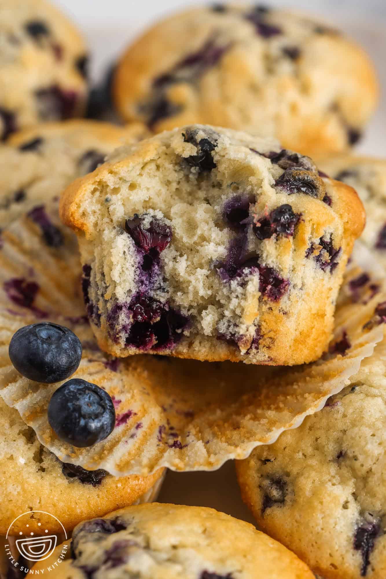 a blueberry muffin that has had the paper peeled away and a bite taken, on top of other blueberry muffins.
