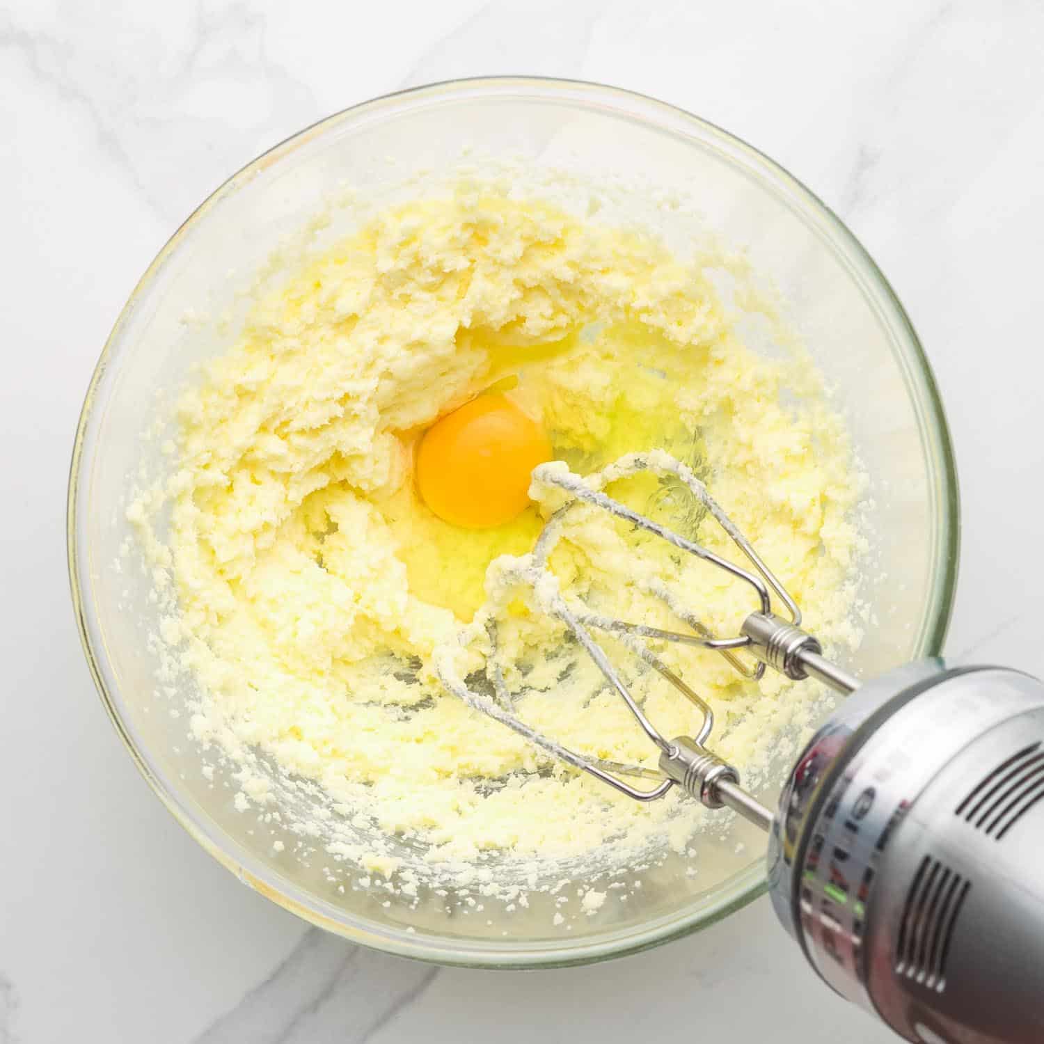 creamed butter and sugar in a glass bowl. An egg is being mixed with with a handheld electric mixer.