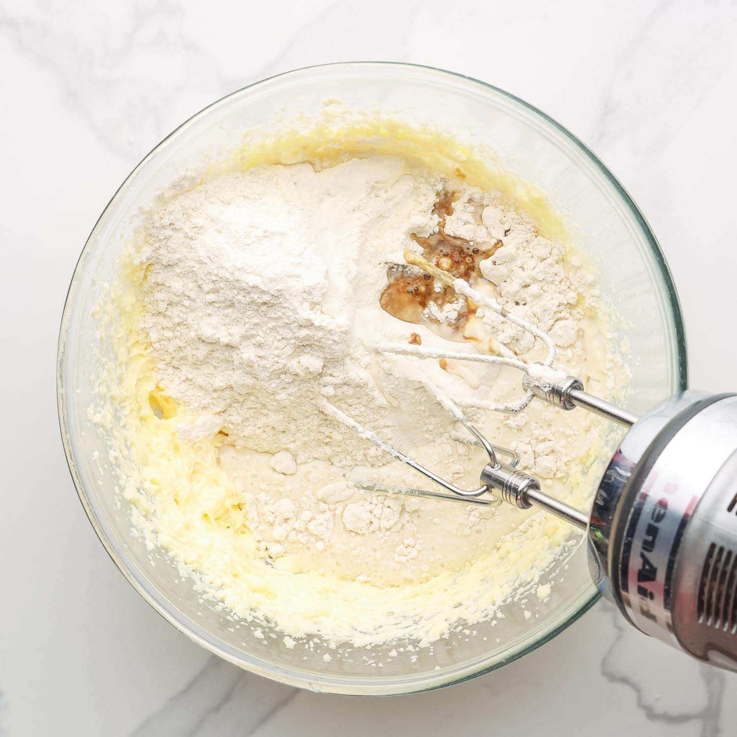 flour added to wet ingredients in a glass mixing bowl, a mixer is combining the ingredients to make muffins.
