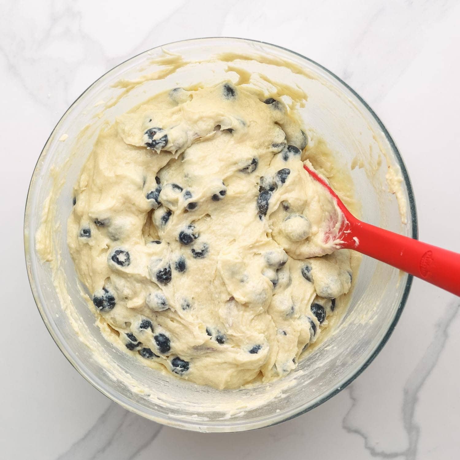 blueberry muffin batter in a glass bowl stirred with a red spatula.
