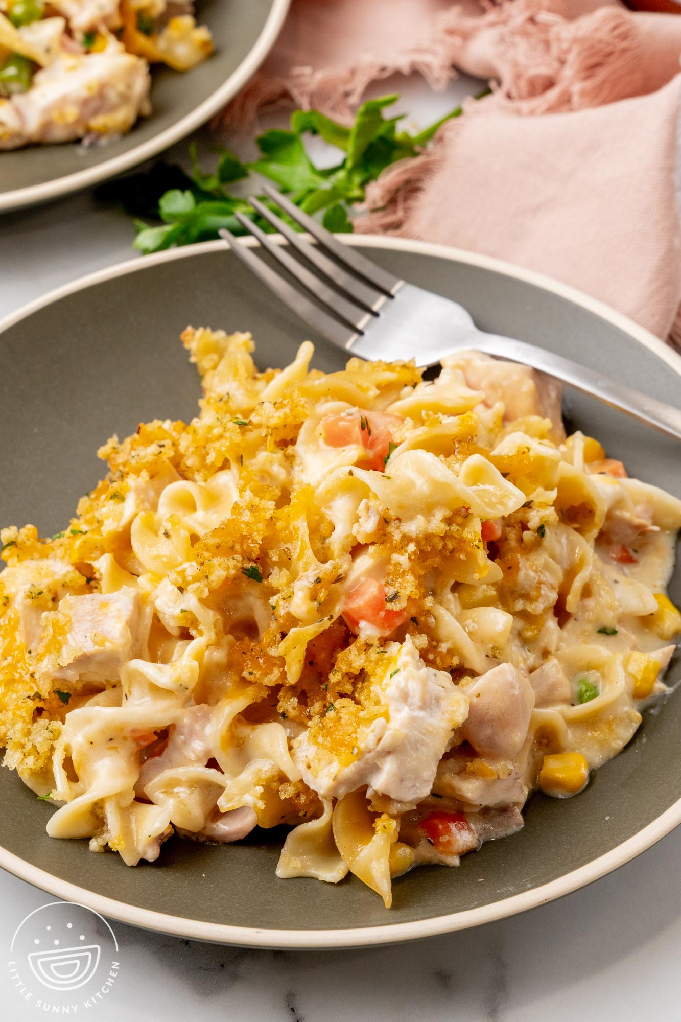 A large serving of homemade chicken noodle casserole on a gray plate with a fork. in the background is a light pink napkin.