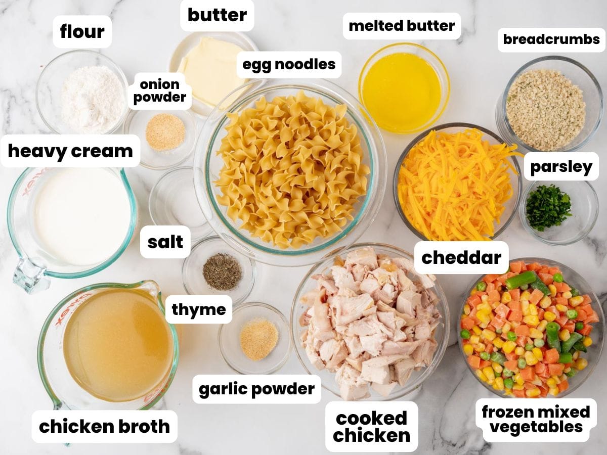 Ingredients needed to make chicken noodle casserole, measured and arranged in glass bowls on a marble surface. Included are egg noodles, mixed veggies, cooked, cubed chicken, shredded cheese, heavy cream, chicken broth, seasonings.