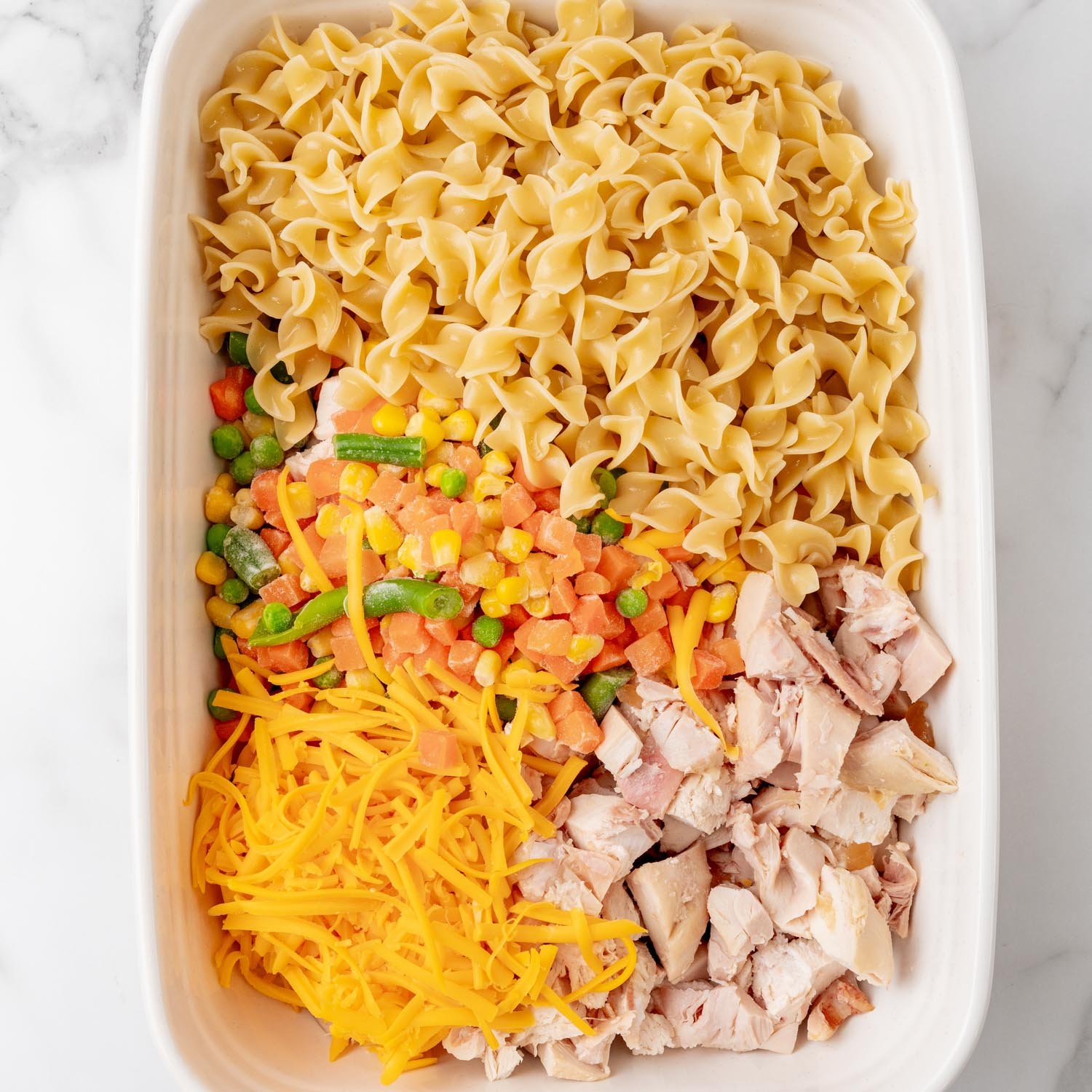 Egg noodles, cooked chicken, frozen mixed vegetables, and shredded cheese in a white rectangular casserole dish.